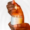 Mão segurando frasco aberto do HAMI Creme Hidratante da Nutri Bu Alma, com espátula a retirar o produto.